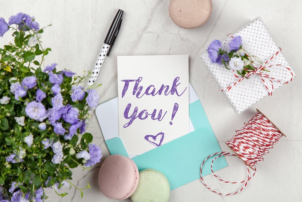A beautifully arranged thank you card with lavender flowers, macarons, and gift package on a marble desk.